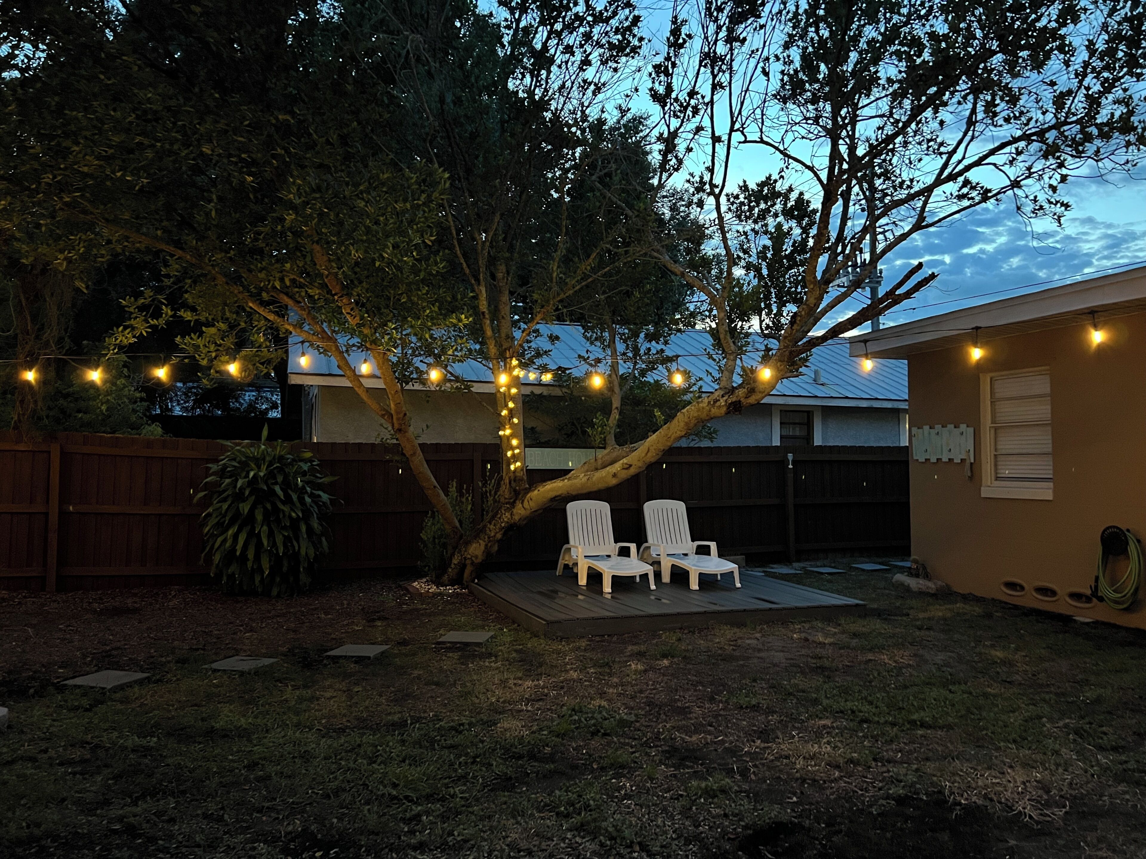 A backyard with a tree, two white chairs, and string lights.