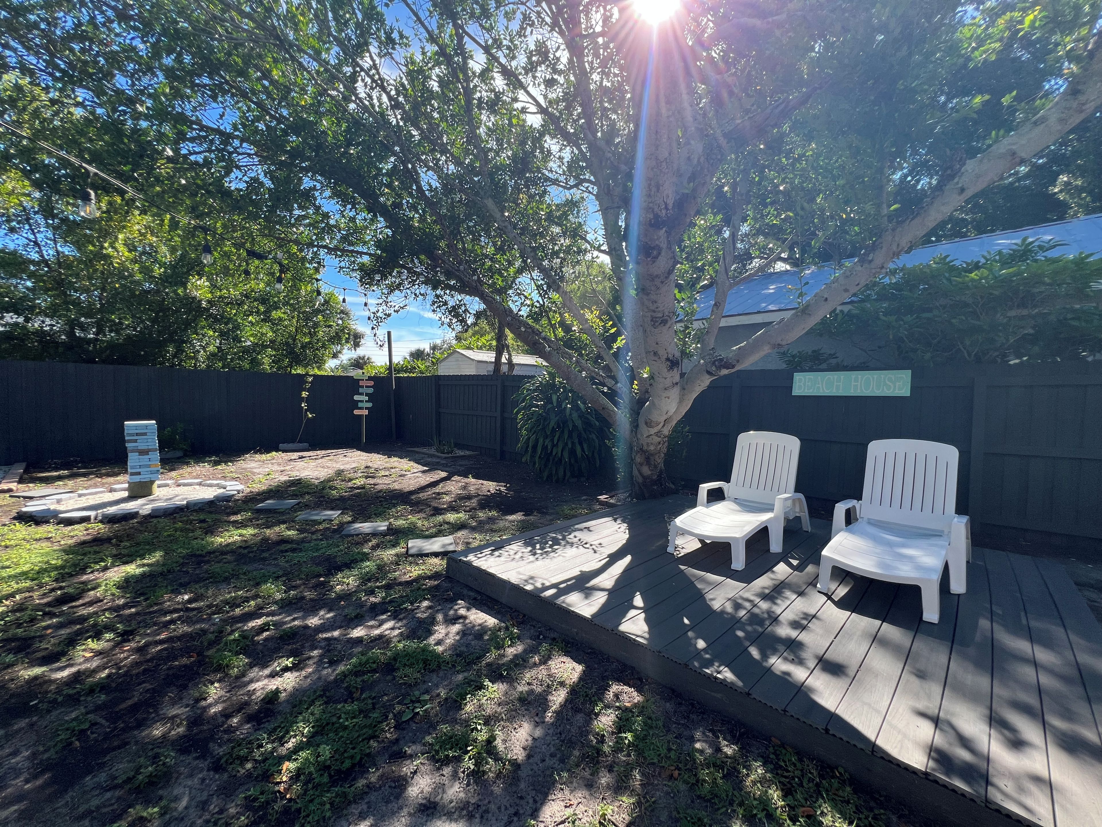 A backyard with a wooden deck, two white chairs, a stone pathway, and a sign reading 'BEACH HOUSE'.