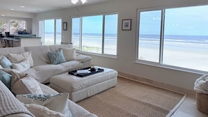 A spacious living room with a large window overlooking a beach, a ceiling fan, and a kitchen area visible through the window.