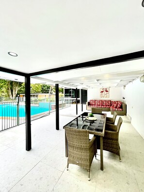 An outdoor patio area with a dining table and chairs, a pool in the background, and a red sofa. - Spacious Hideaway Retreat, Pool , Spa,  Acreage, Billiards, 7 Bedrooms, Brisbane (Brookfield)