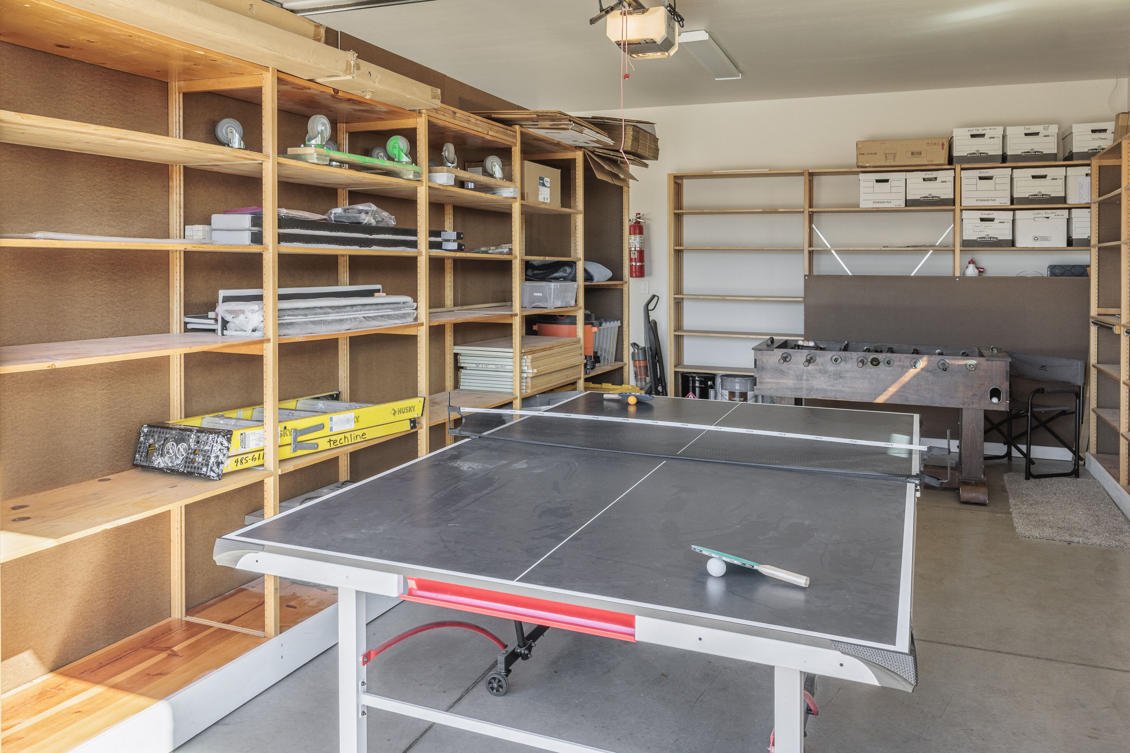A room with a ping pong table, shelves with boxes, and a foosball table.