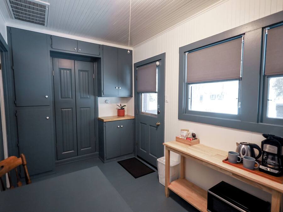 A compact kitchen with grey cabinets, a wooden table with a coffee maker, and a window with blinds.