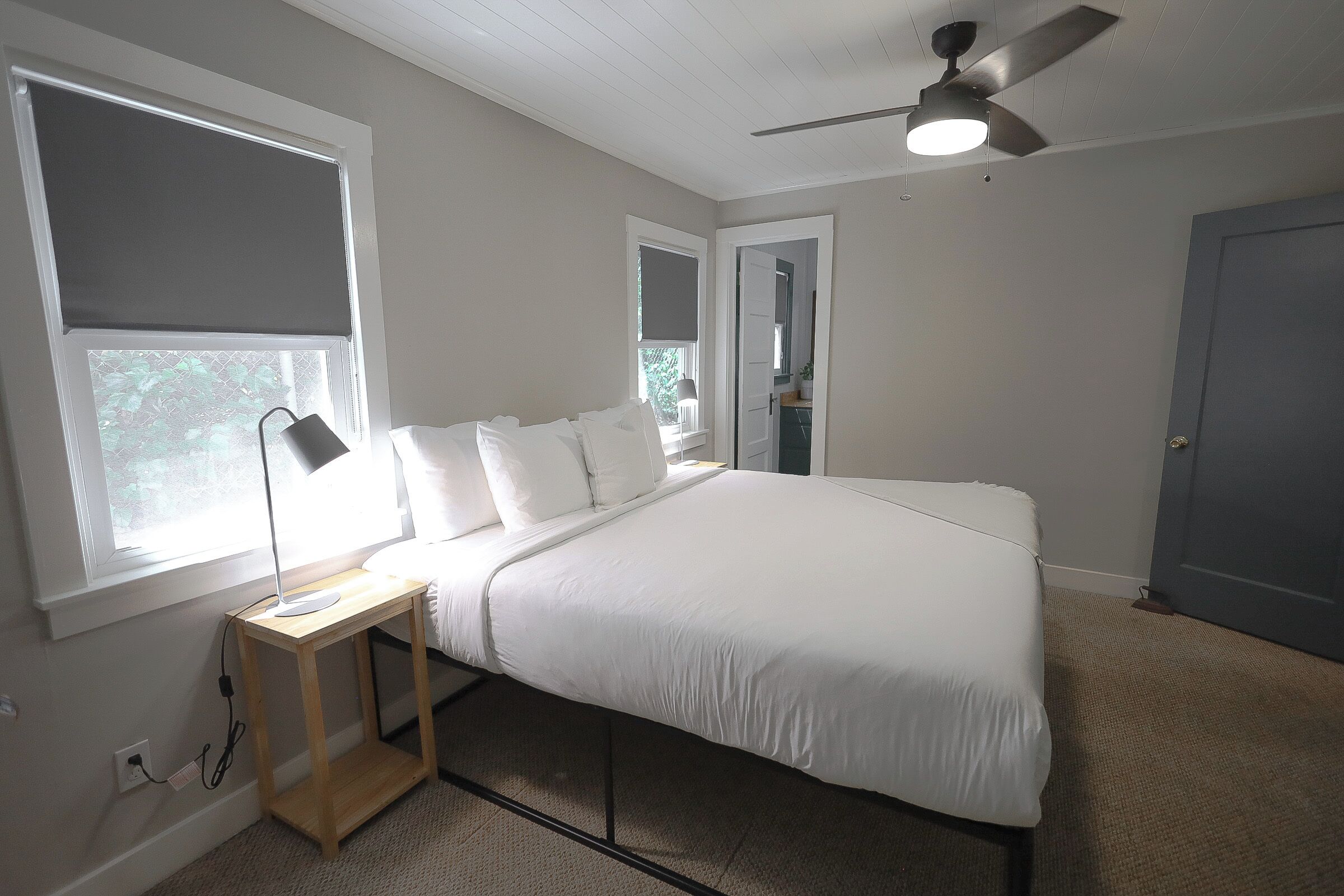 A bedroom with a bed, a desk with a lamp, a ceiling fan, and a window with blinds.