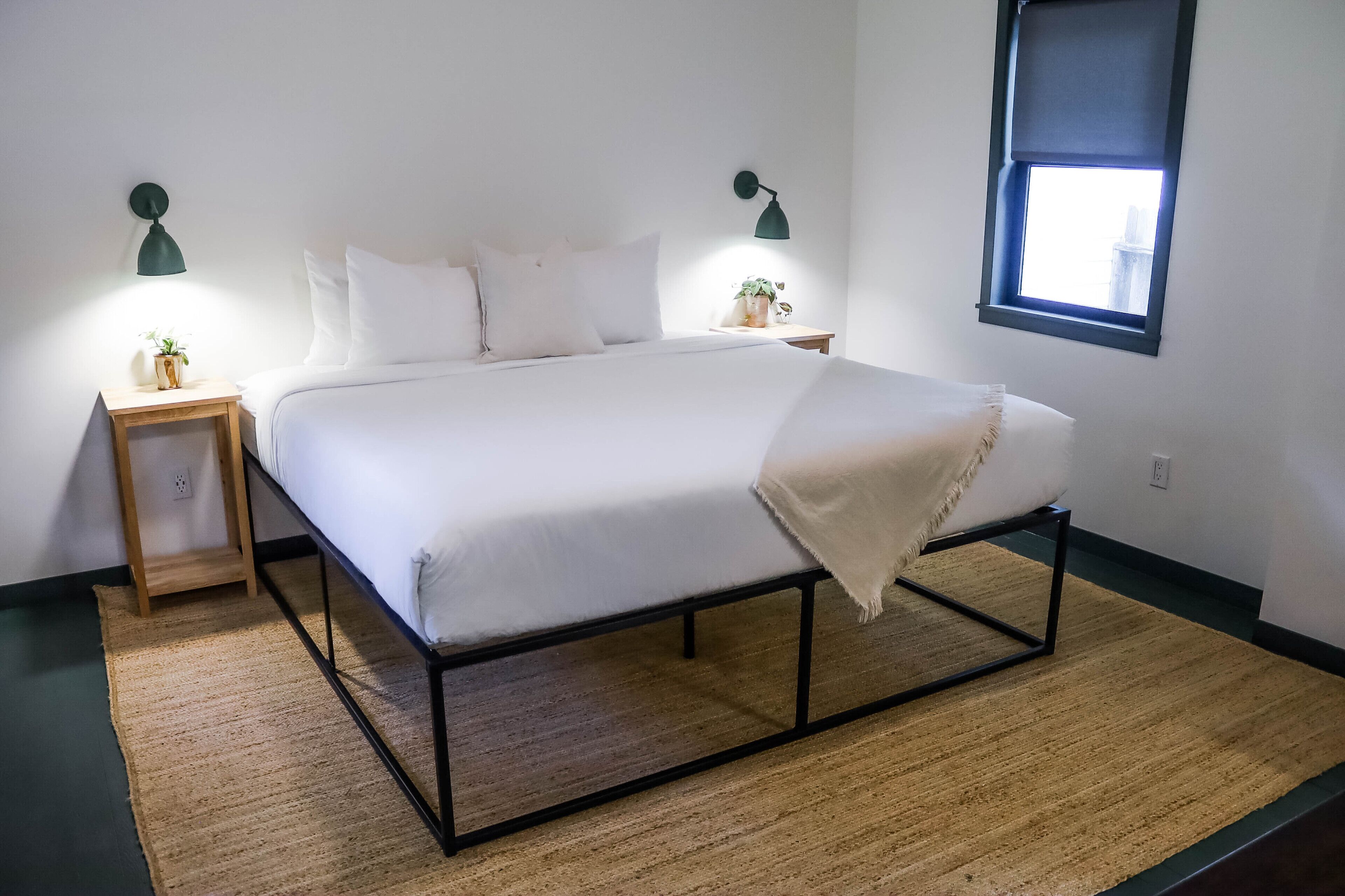 Un dormitorio ordenado con cama, mesitas de noche, una planta pequeña y una ventana con vistas.
