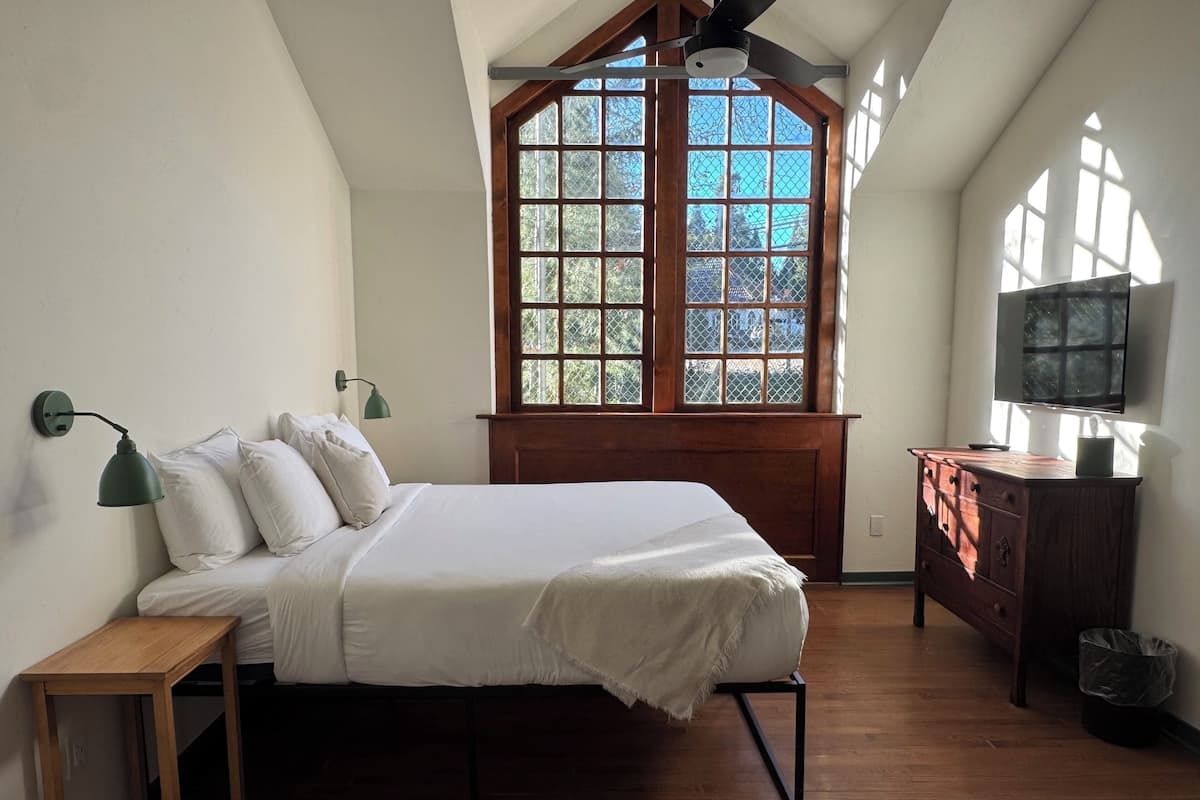 A bedroom with a bed, a wooden dresser, a TV, and a window with a view of trees.