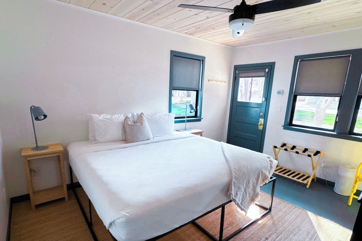 A bedroom with a bed, a bedside table, a lamp, a window, and a door.