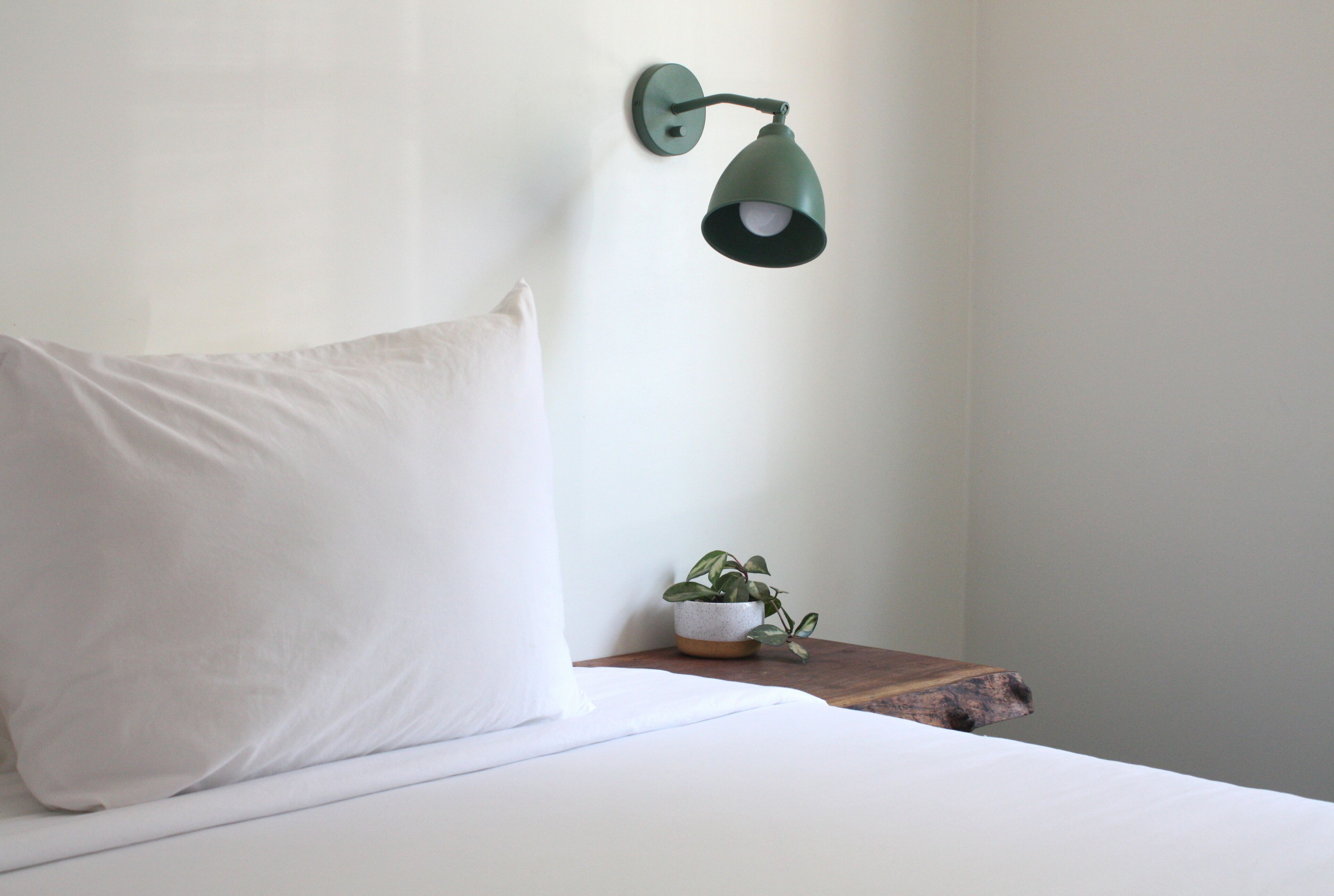 Cama bem arrumada com travesseiro branco e luminária verde fixada na parede ao lado.