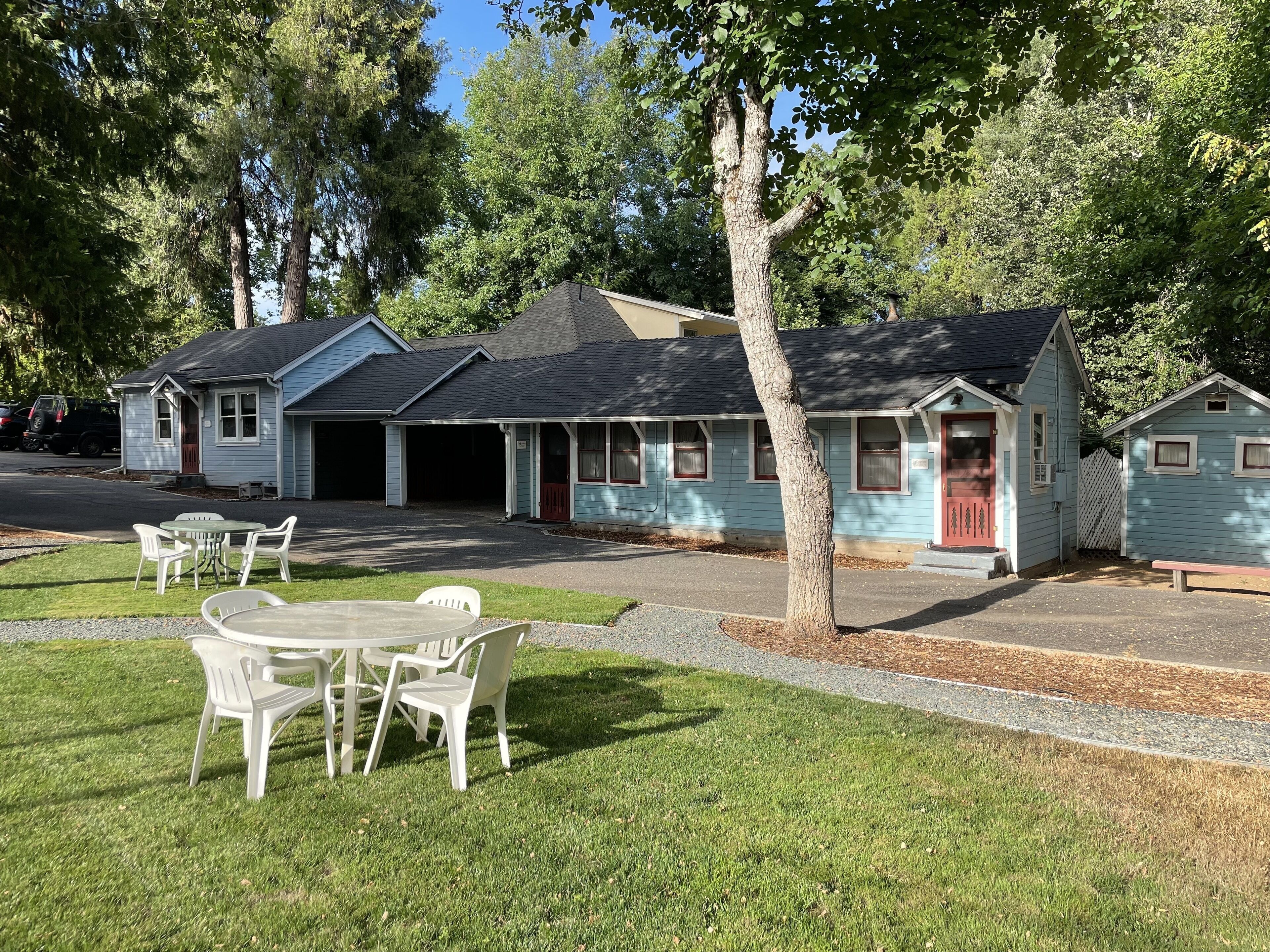 A row of light blue cabins with a small table and chairs in front, surrounded by trees.
