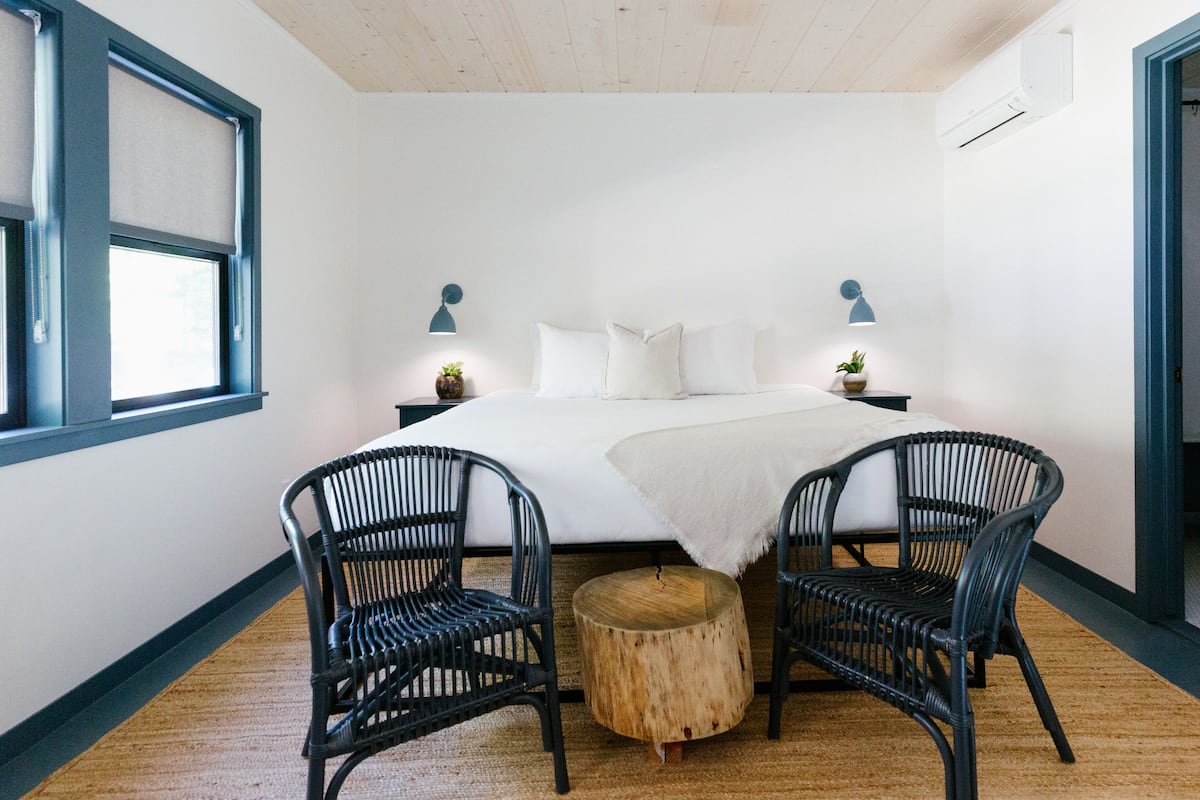 A bedroom with a bed, two nightstands with lamps, two wicker chairs, a round wooden stool, and a window with a roller blind.