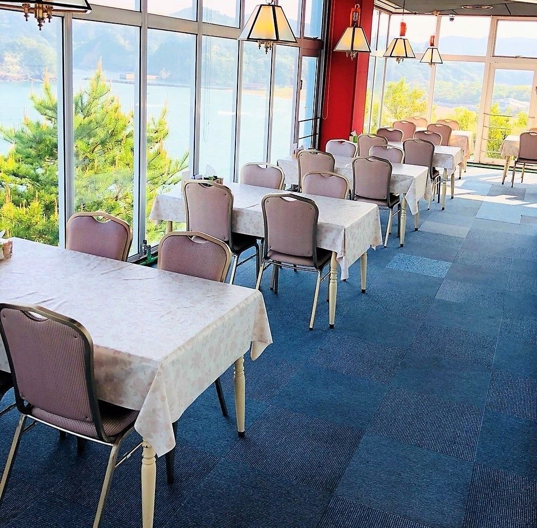 A restaurant with tables and chairs arranged in rows, large windows offering a view of trees and a body of water.