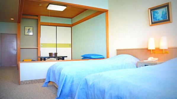 Quarto de hotel com duas camas, uma escrivaninha, uma cadeira, uma mesinha com um bule, um travesseiro azul, um quadro na parede e uma mesa de cabeceira com abajur.