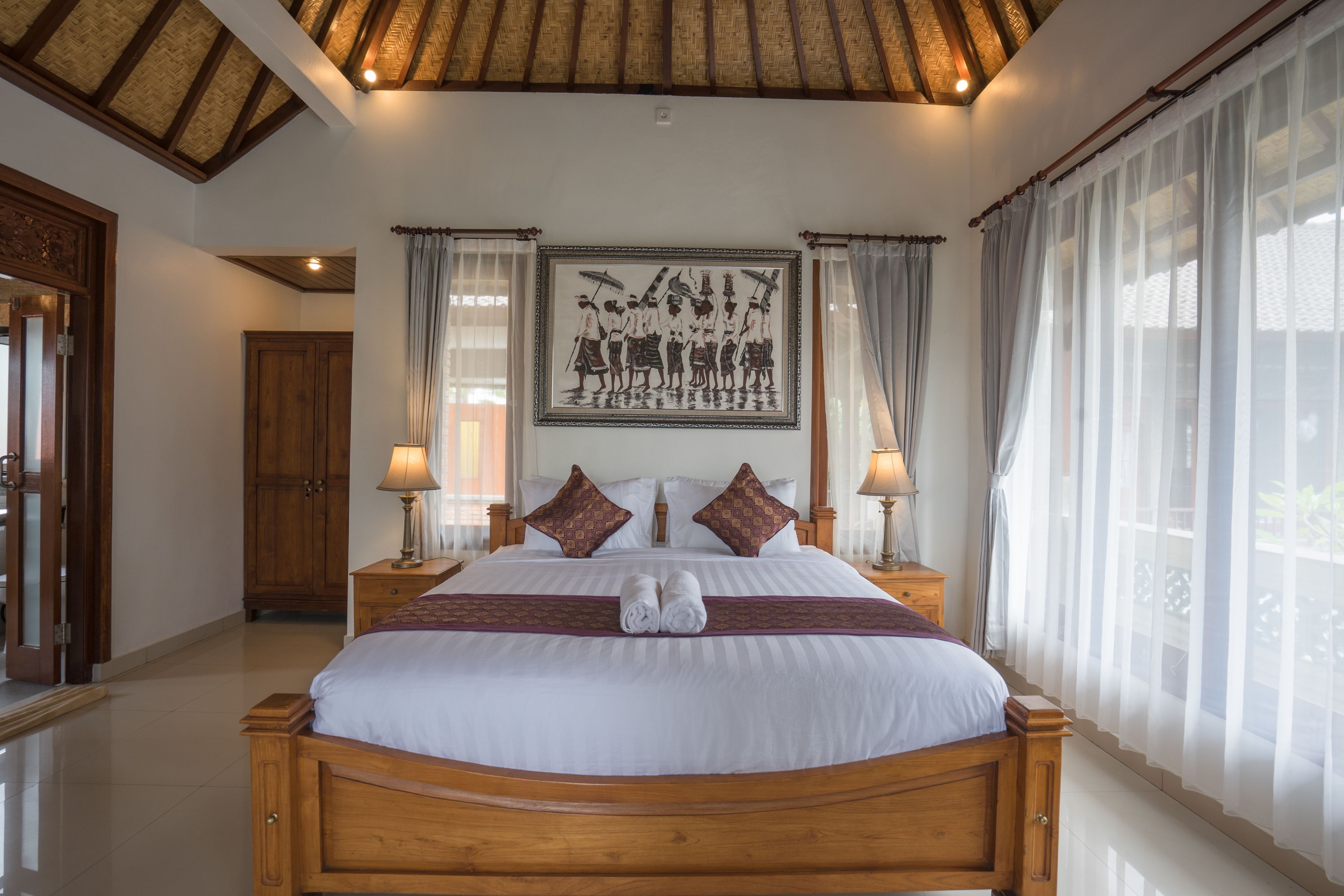 A large bed with a wooden frame, two bedside tables with lamps, a framed picture above the bed, and sheer curtains on the windows.