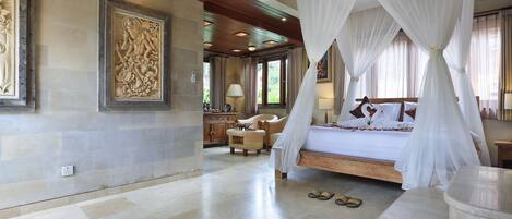 A bedroom with a four-poster bed, a sitting area with a chair and ottoman, and artwork on the walls.