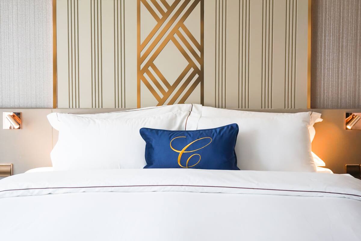 A neatly made bed with white linens and a single blue pillow with a gold letter 'C'.