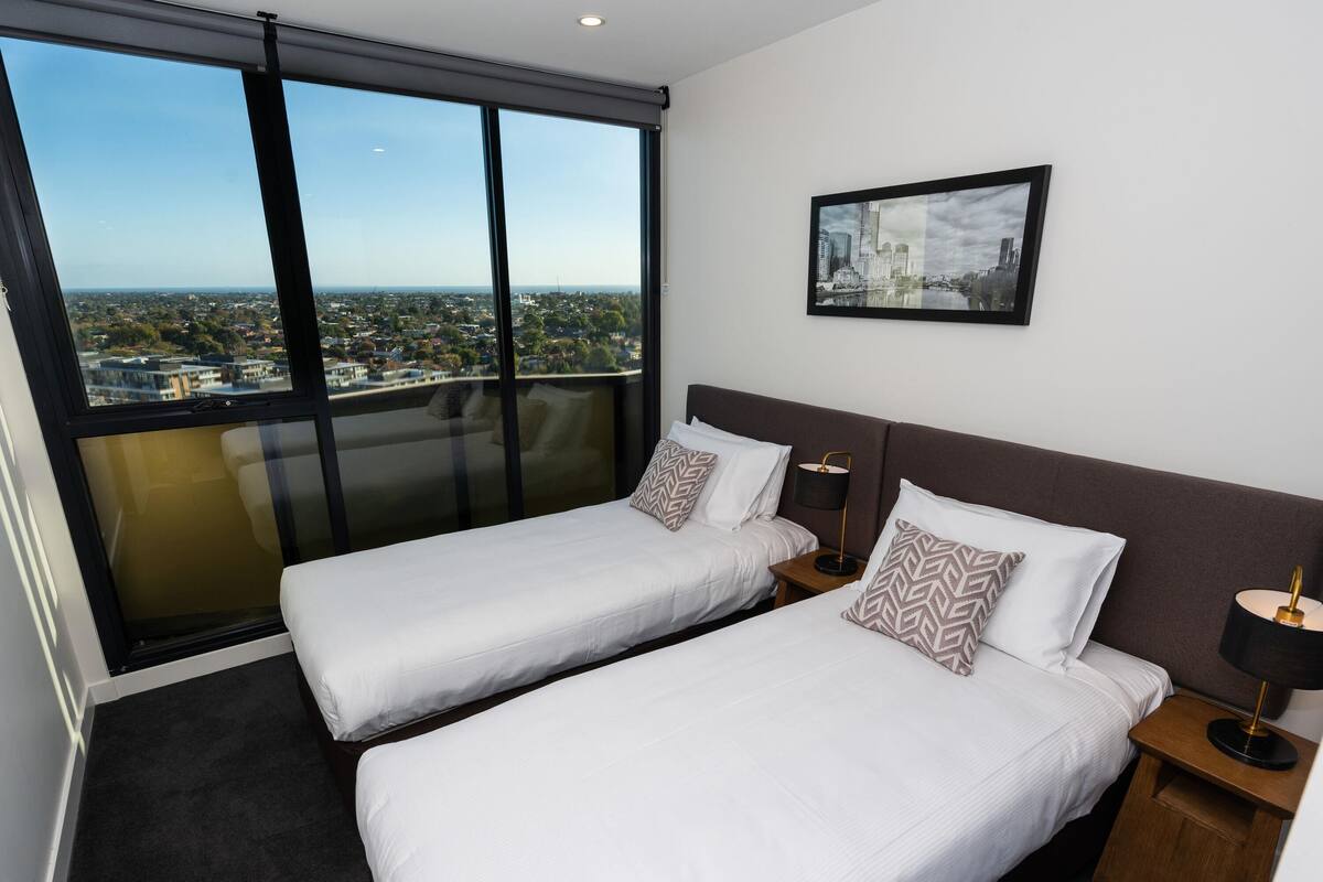 A hotel room with two beds, large windows, a city view, and a framed picture on the wall.