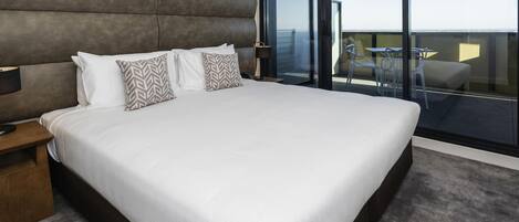 A modern hotel room with a large bed, a nightstand, a desk, and a balcony with a table and chairs.