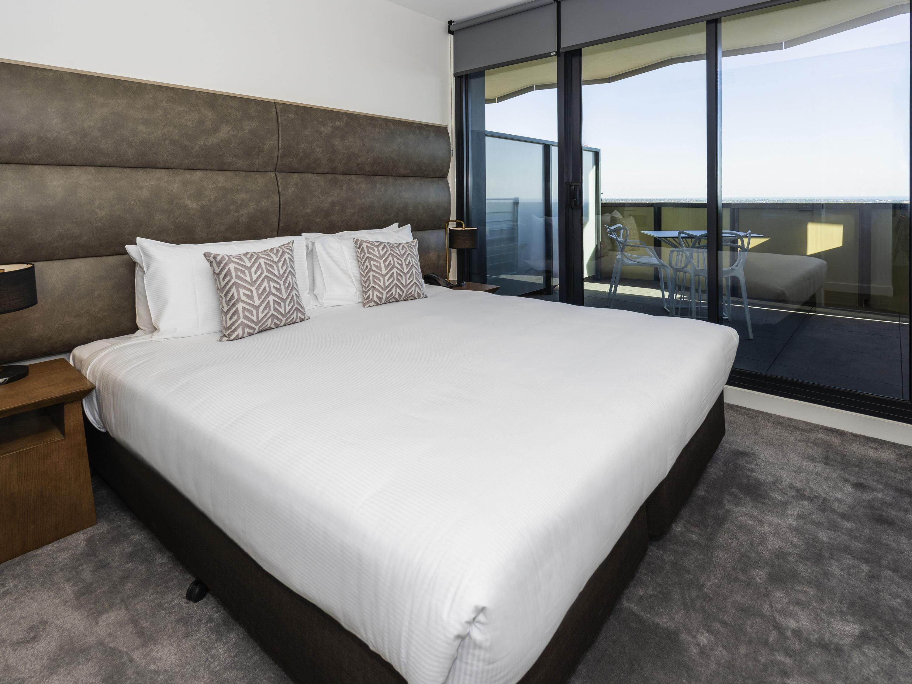 A modern hotel room with a large bed, a nightstand, a desk, and a balcony with a table and chairs.