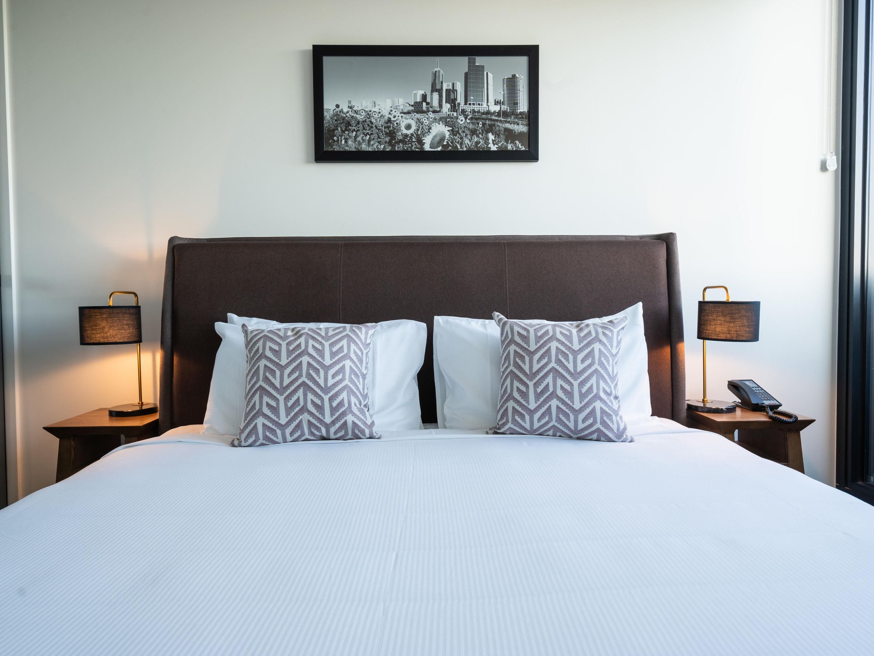 A hotel room with a large bed, two nightstands, two lamps, a cityscape picture on the wall, and a window.