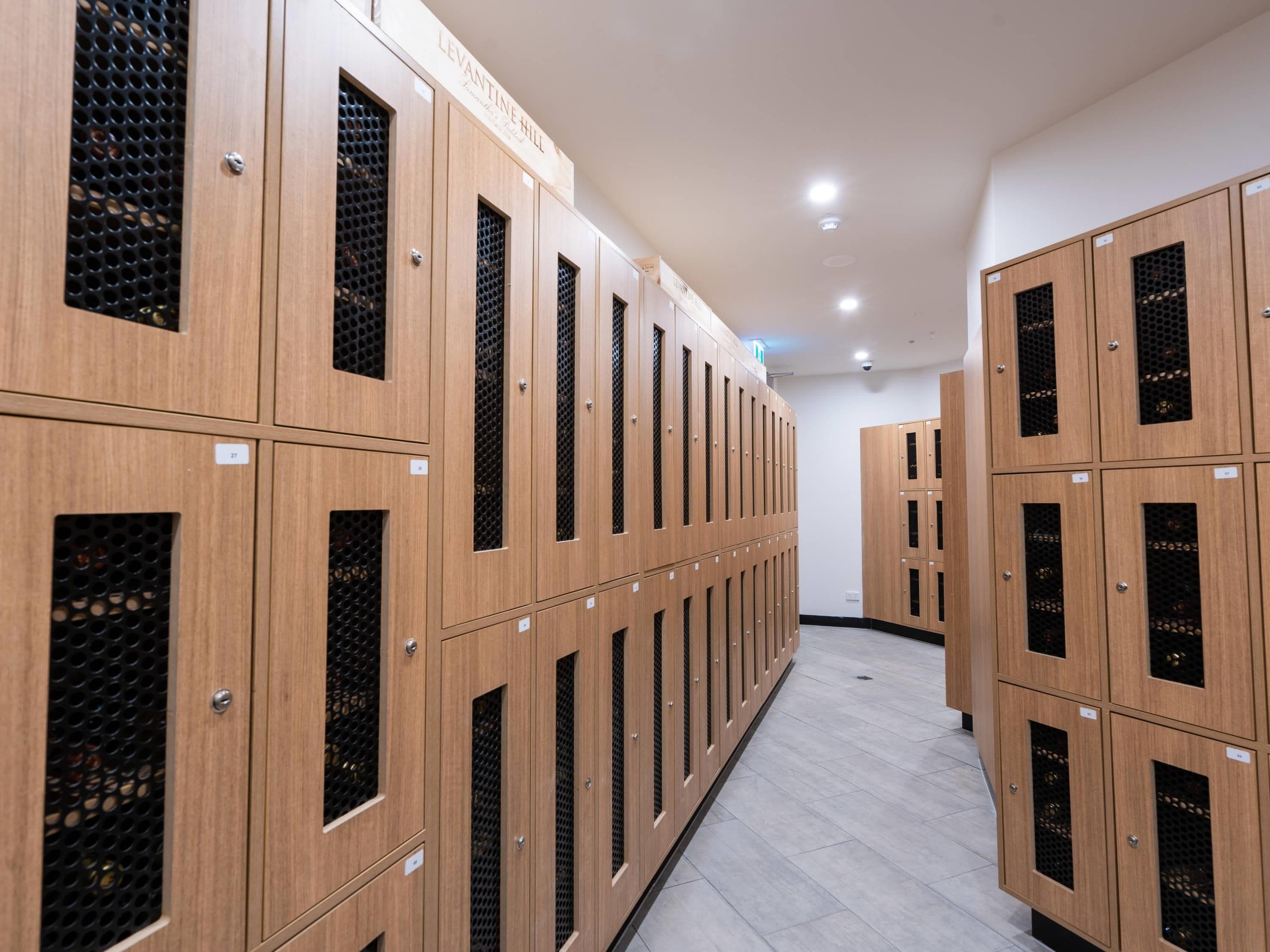 A row of wooden lockers with mesh windows, labeled with numbers, in a hallway with a tiled floor.