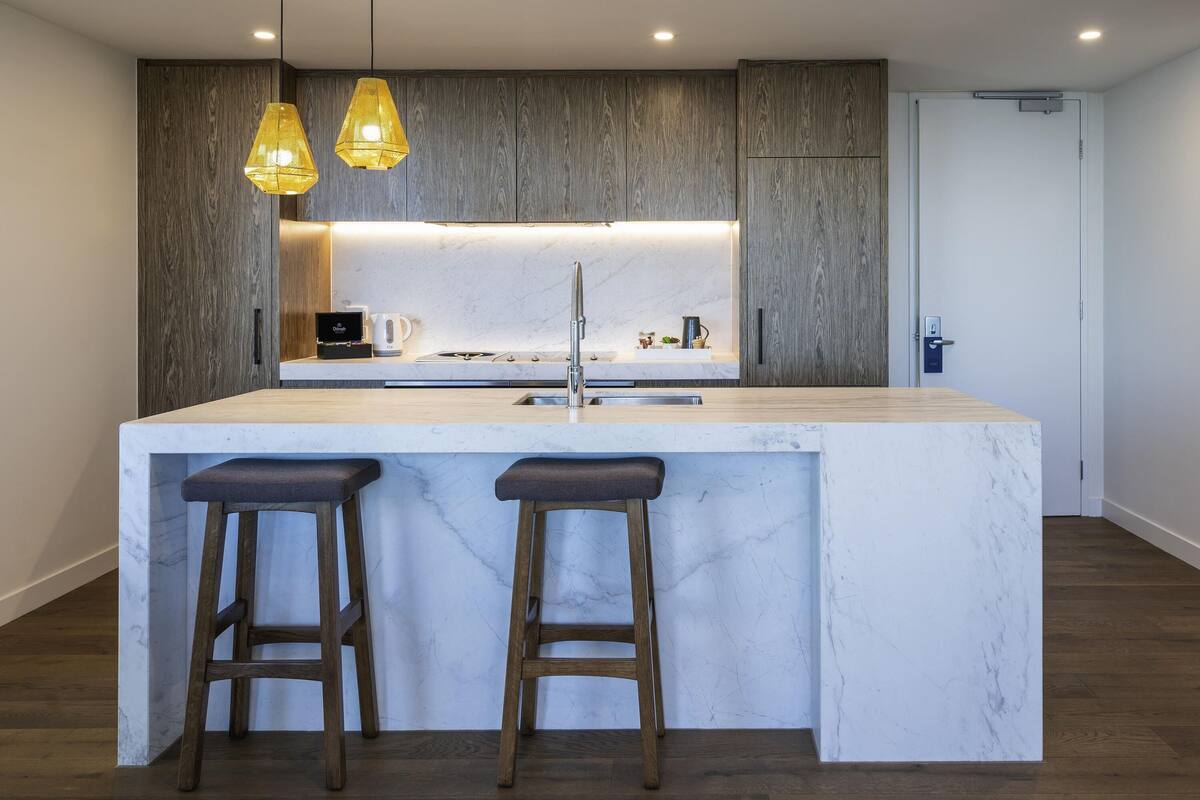 A modern kitchen with a marble island, two bar stools, pendant lights, and wooden cabinetry.