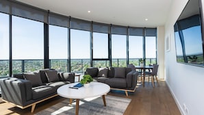 Una sala de estar moderna con ventanas amplias, una mesa redonda blanca, sofás grises y vistas de un paisaje urbano.
