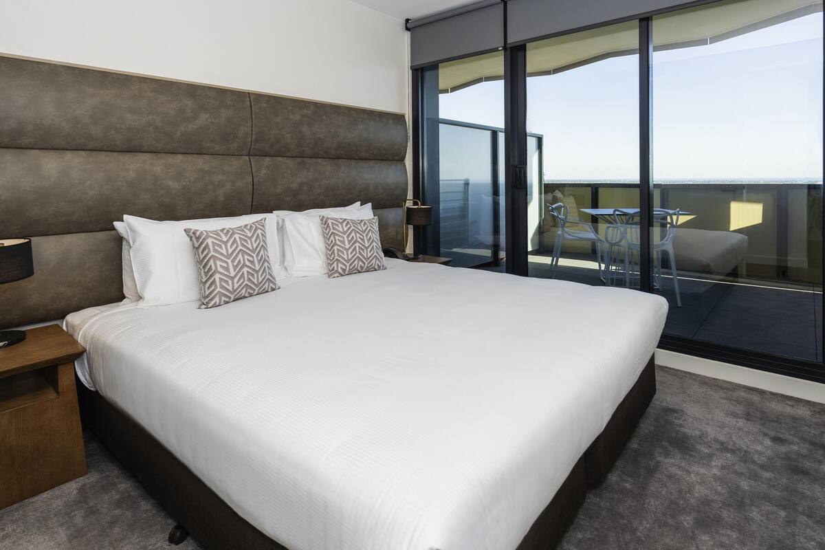 A modern hotel room with a large bed, a nightstand, a desk, and a balcony with a table and chairs.