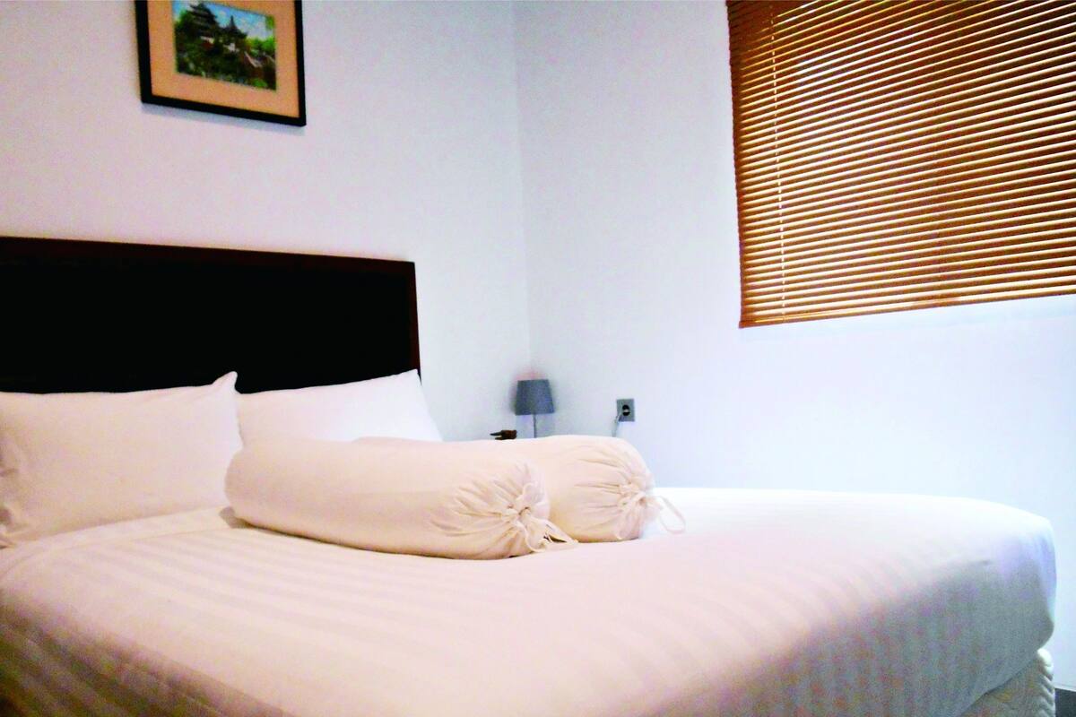 Una cama bien tendida con un cabecero oscuro, una lámpara de noche individual y una ventana con persianas.