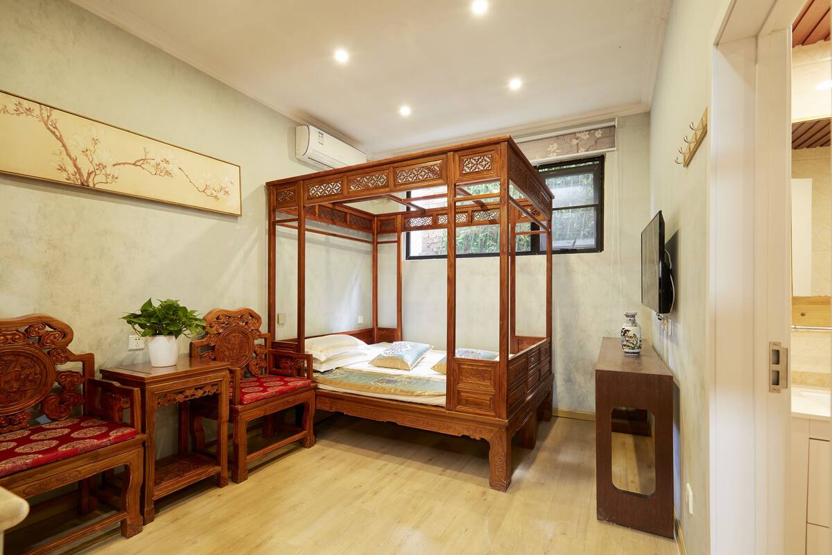 A traditional wooden four-poster bed with a matching nightstand, a small table with a plant, and two wooden chairs with red cushions.