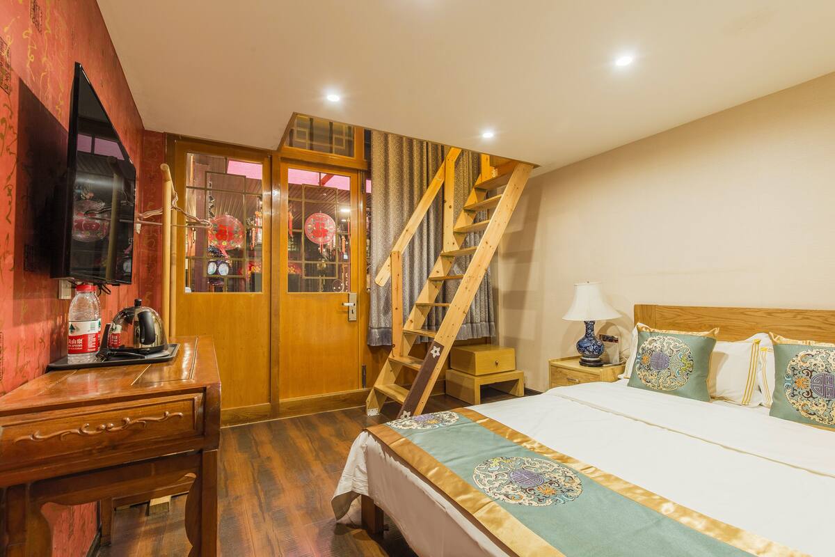 A hotel room with a wooden desk, a bed with patterned bedding, a wooden staircase, and a television on the wall.