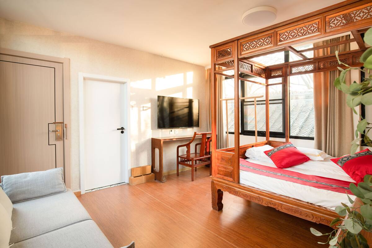 A bedroom with a wooden four-poster bed, a desk with a chair, a television, and a window with curtains.