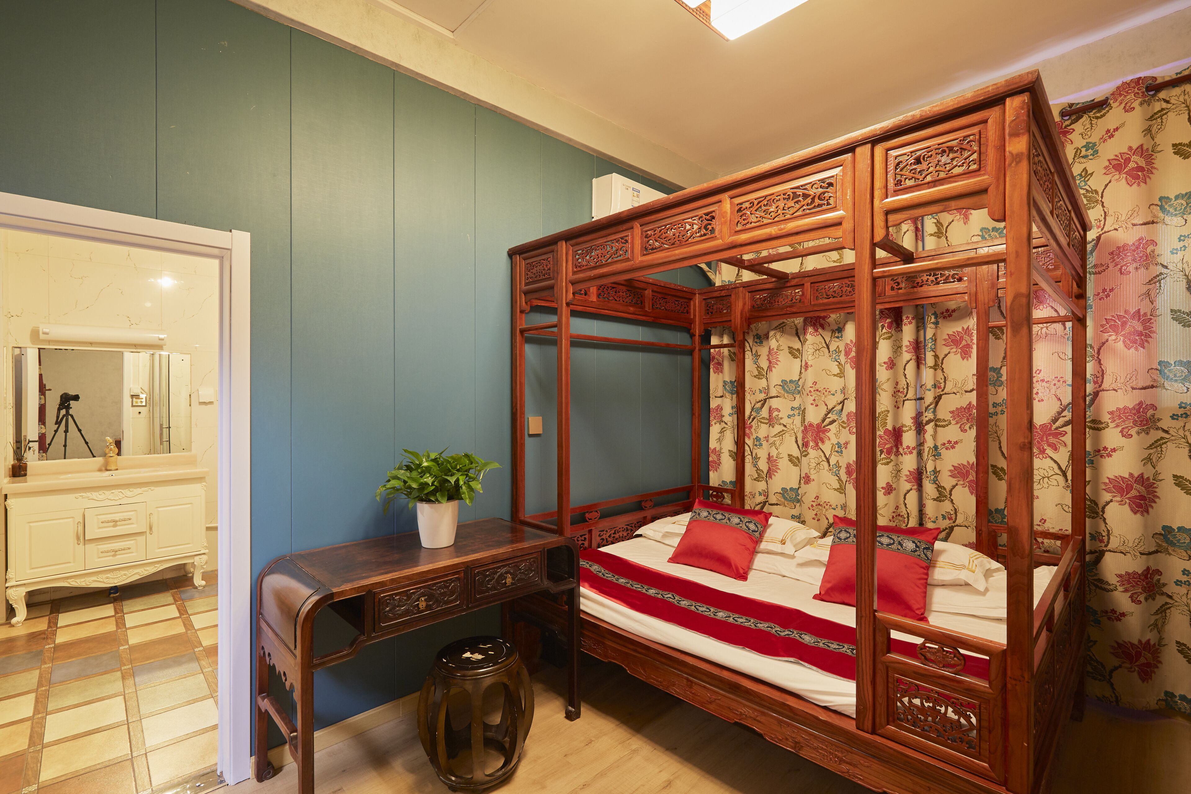 A traditional four-poster bed with intricate carvings, a wooden desk with a plant, and a mirror cabinet.