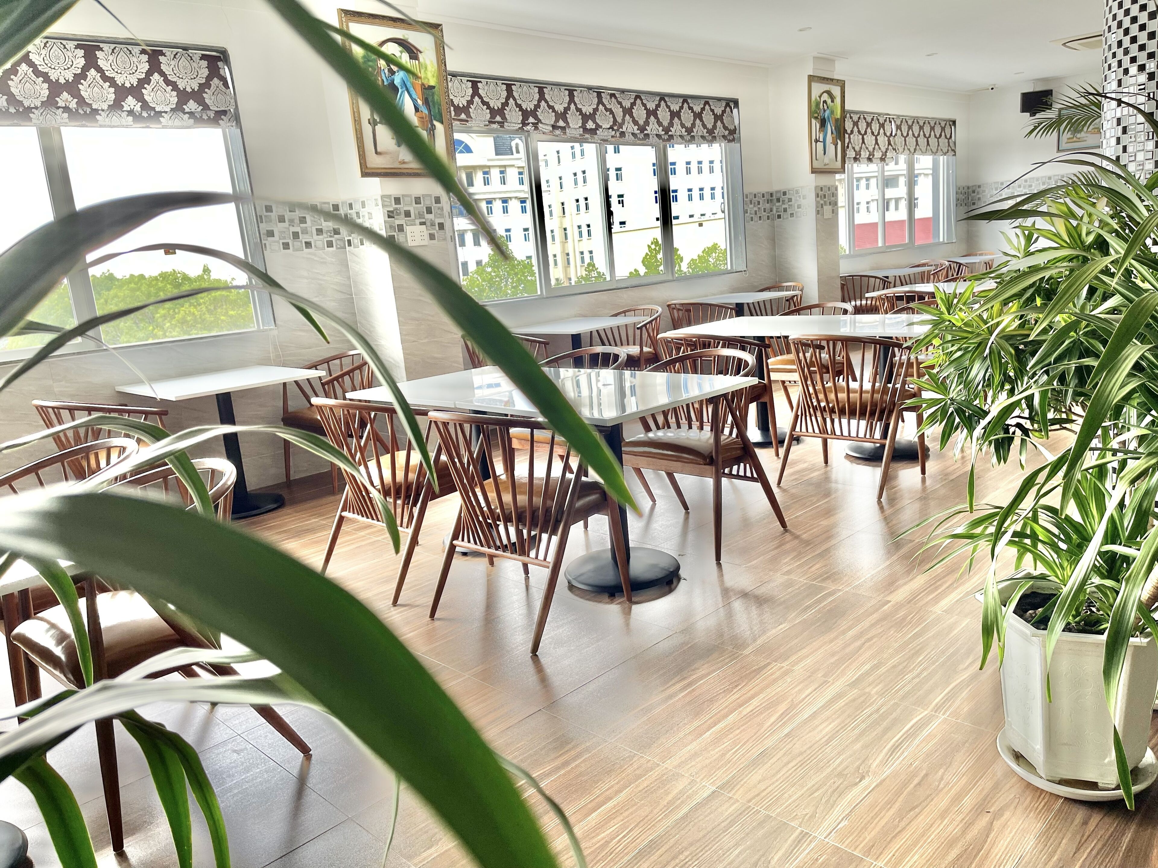 Un restaurante con mesas y sillas de madera, ventanas amplias con cortinas estampadas y plantas en macetas.