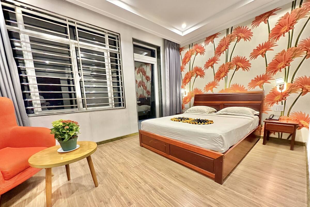 Un dormitorio con una cama de madera, una mesita redonda con una planta, una silla naranja brillante y una ventana con persianas.