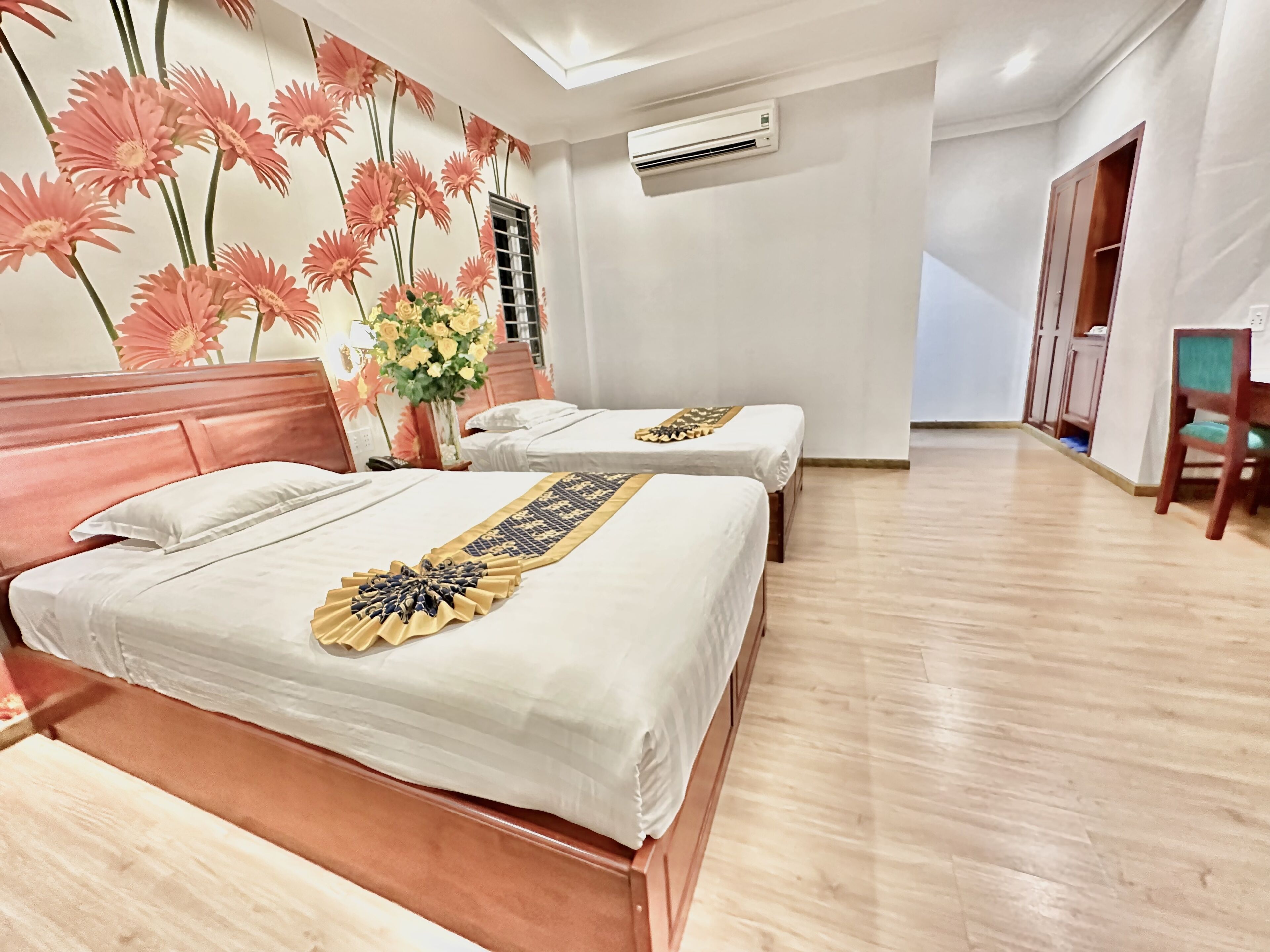 A hotel room with two beds, a wooden floor, a wall with a floral mural, an air conditioning unit, and a wooden wardrobe.