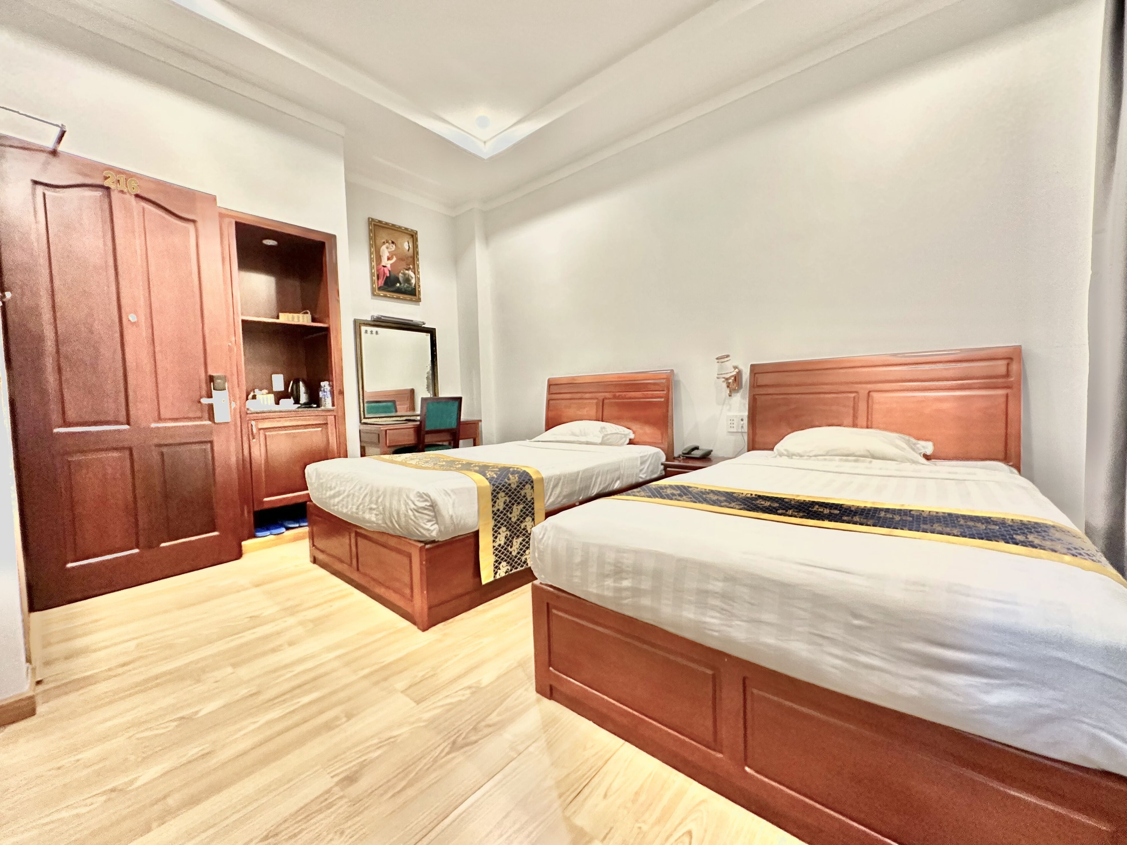 A hotel room with two wooden beds, a wardrobe, a desk with a chair, and a mirror on the wall.