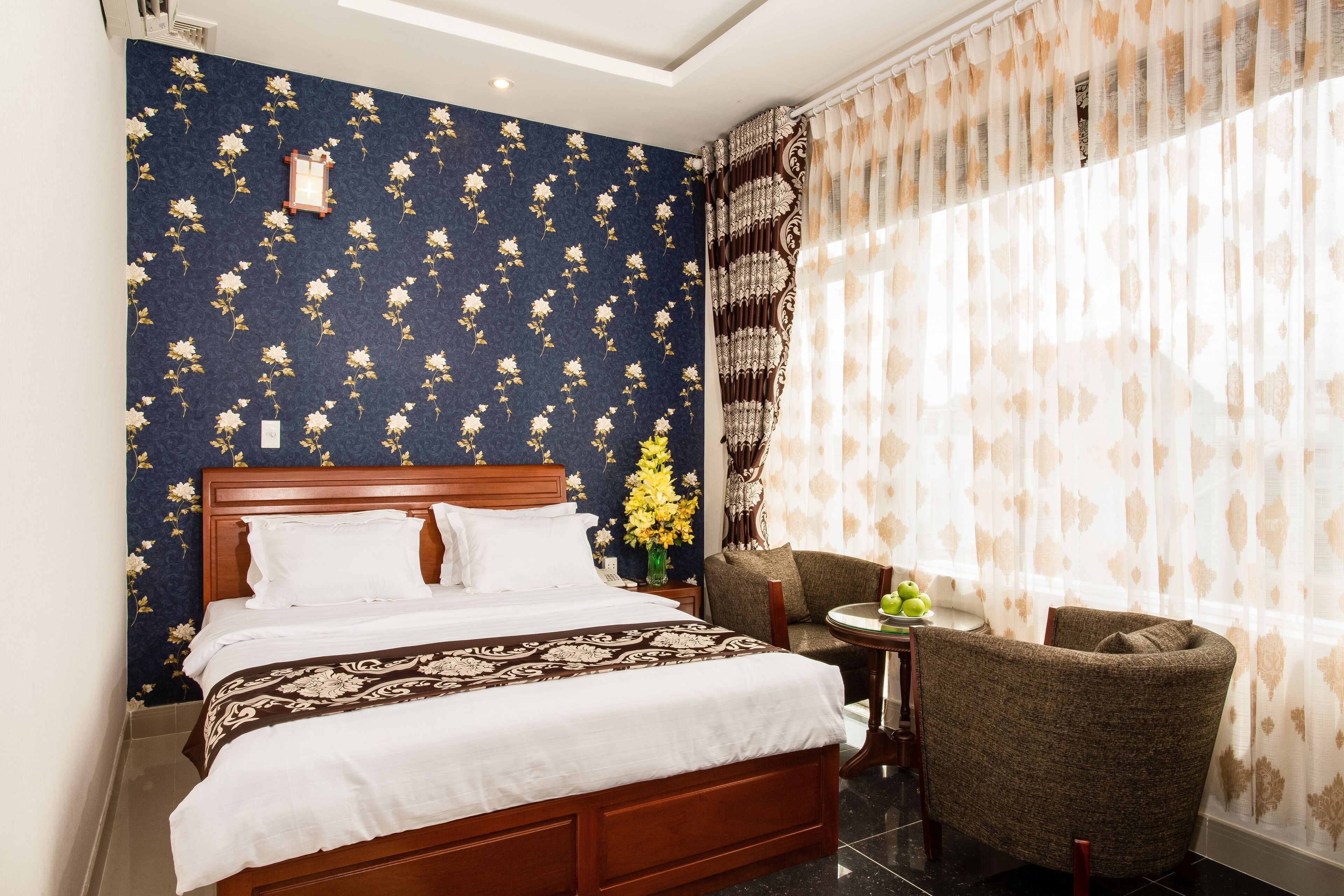 A hotel room with a large bed, a small round table with a chair, a vase with flowers, and a window with curtains.