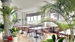 A spacious dining area with wooden flooring, multiple tables, and chairs. There are several potted plants and a large column with a checkered pattern. - Long Bao Chau Hotel (Ho Chi Minh City)