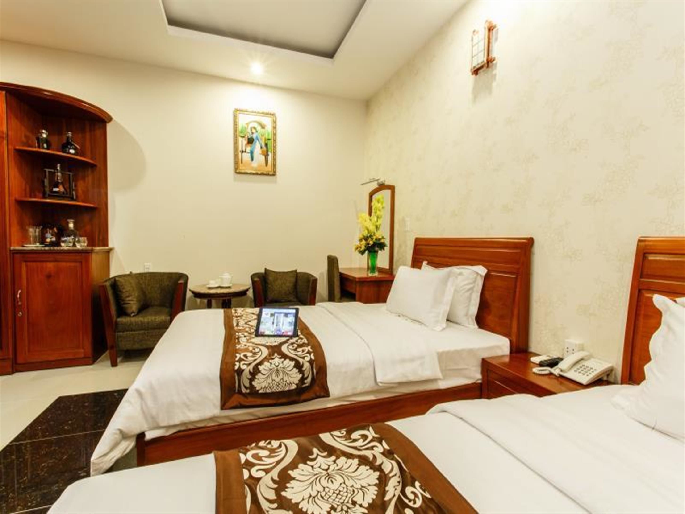A hotel room with two beds, a wooden wardrobe, a small table with a chair, a bedside table with a phone, and a painting on the wall.