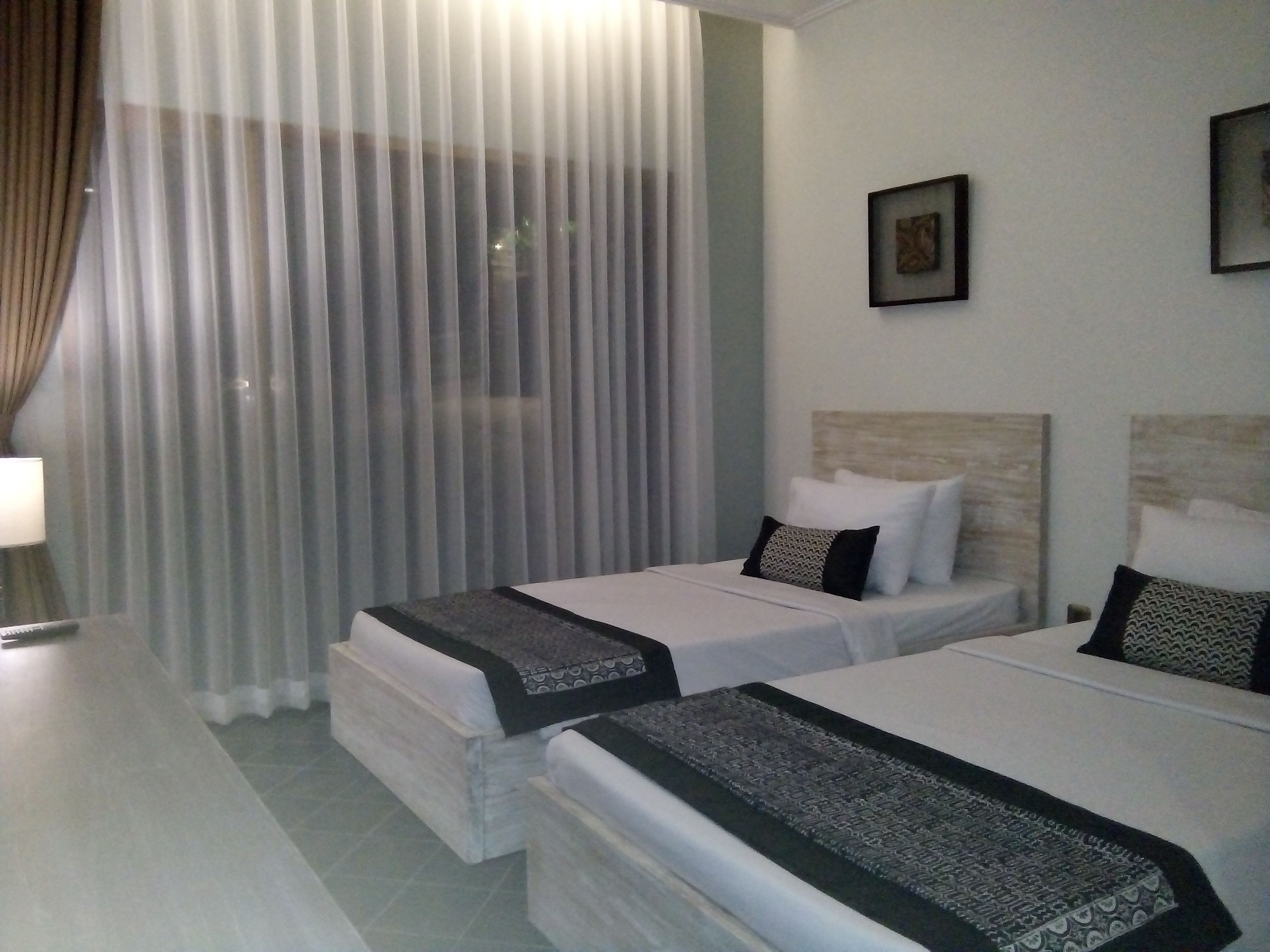 A hotel room with two beds, a large window with sheer curtains, a bedside table with a lamp, and a framed picture on the wall.