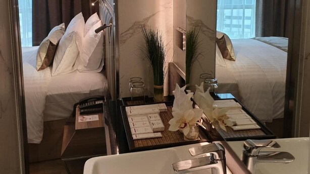 Quarto de hotel com uma cama grande, área de estar separada com mesa e cadeiras, um espelho grande e banheiro com banheira e pia.