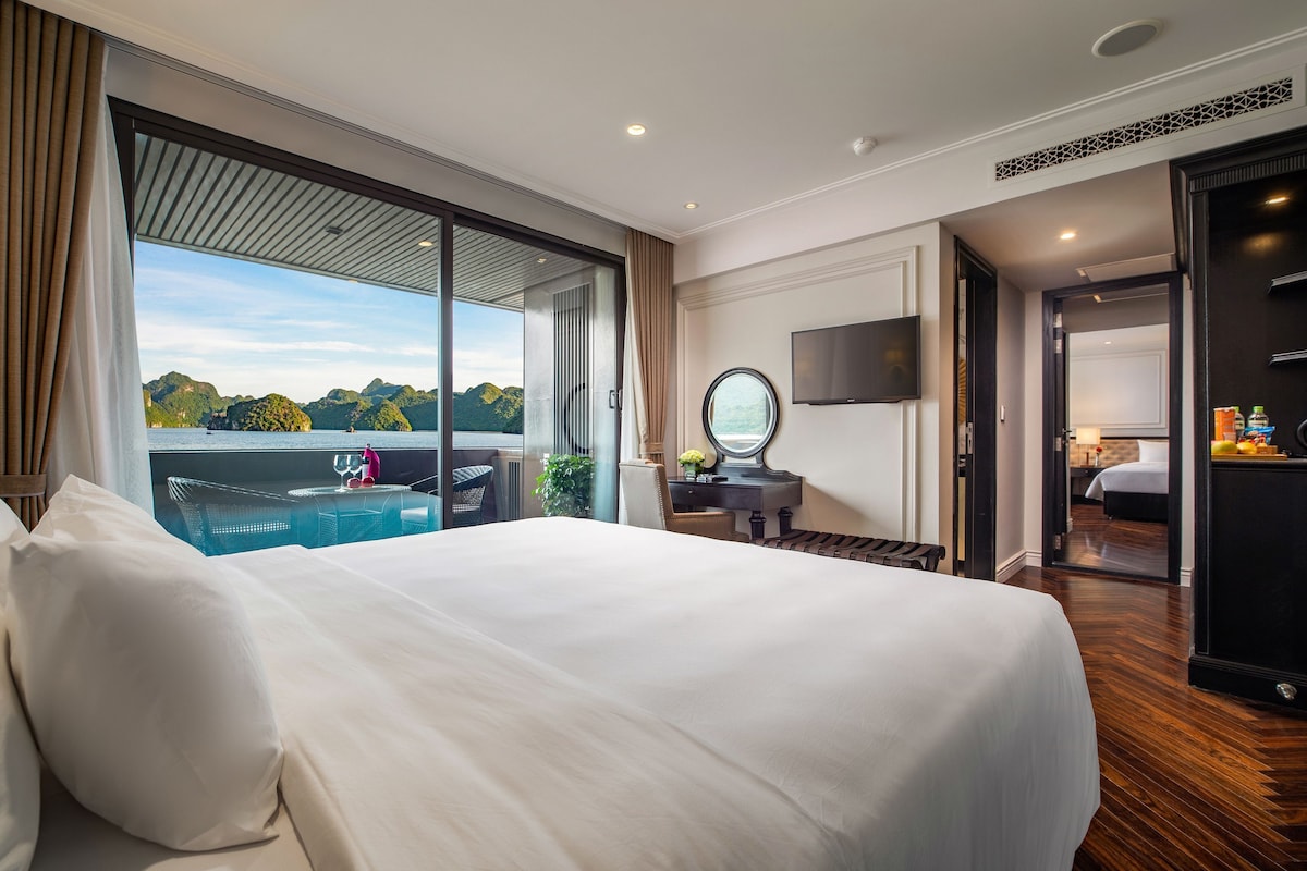 A hotel room with a large bed, a TV, a desk with a mirror, and a view of a body of water and mountains.
