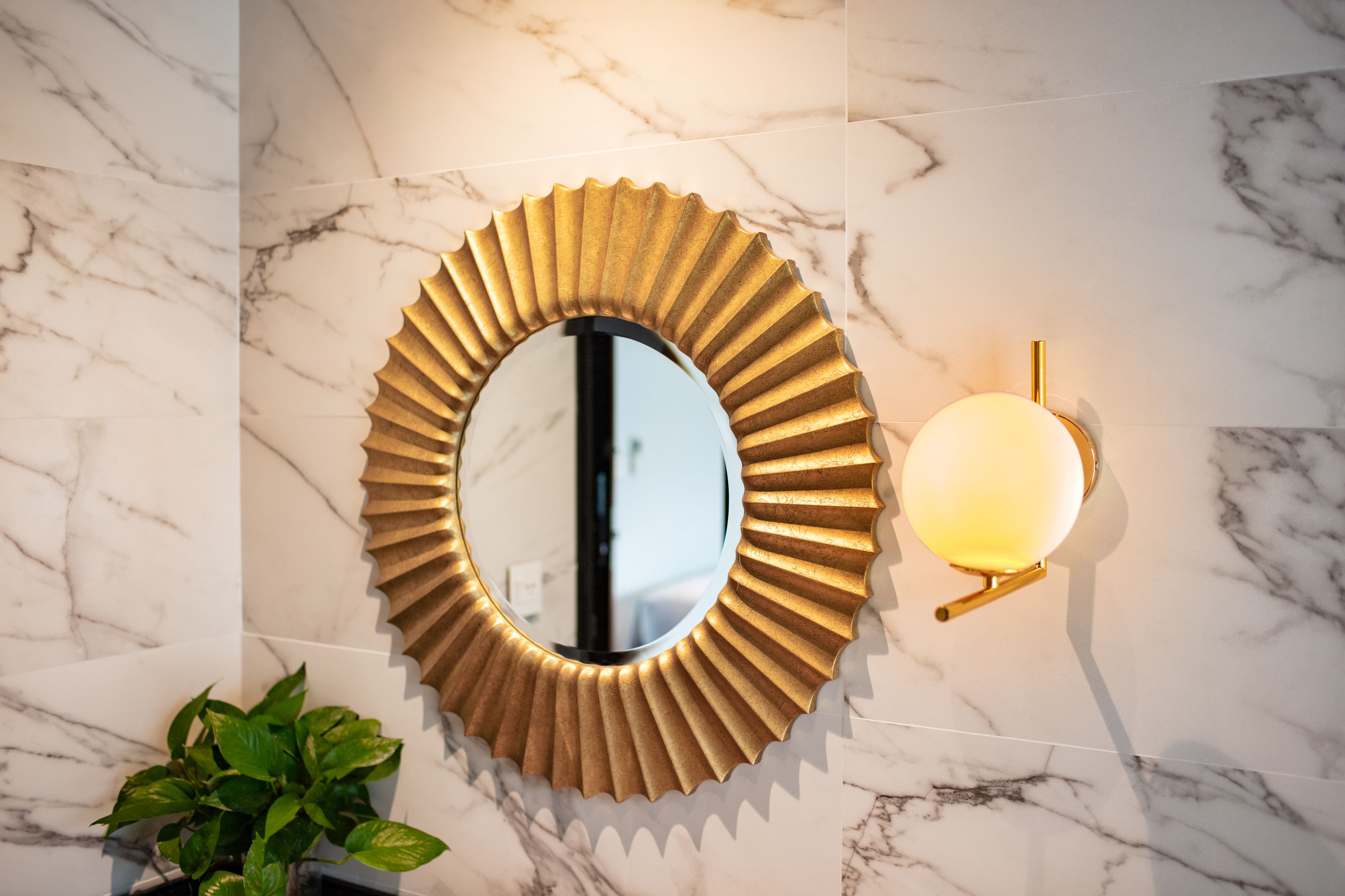 A bathroom with a marble wall, a gold-framed mirror, and a wall-mounted light fixture.