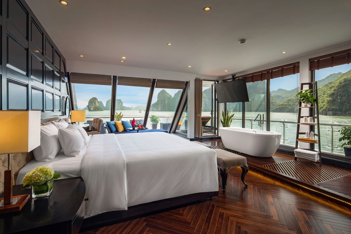 A modern hotel room with a large bed, a bathtub, and a view of a body of water and mountains.