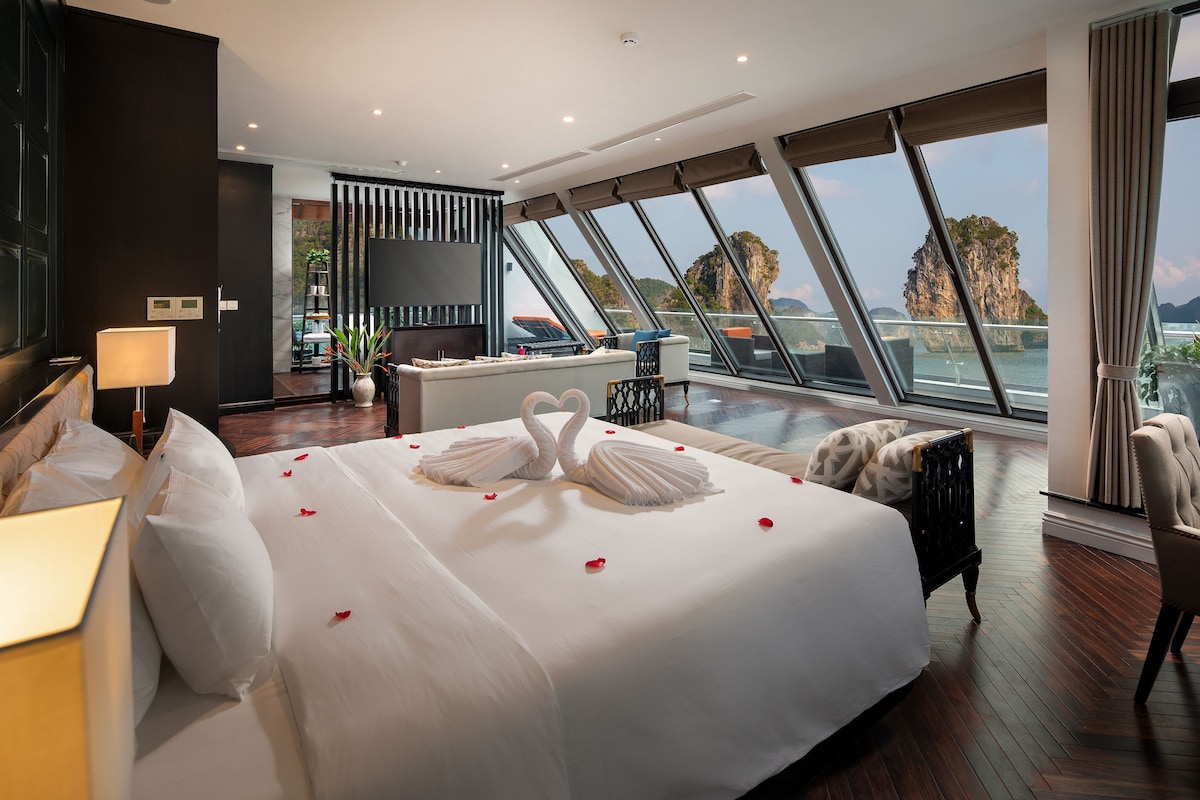 A hotel room with a large bed, a desk, a chair, and a view of the sea through large windows.