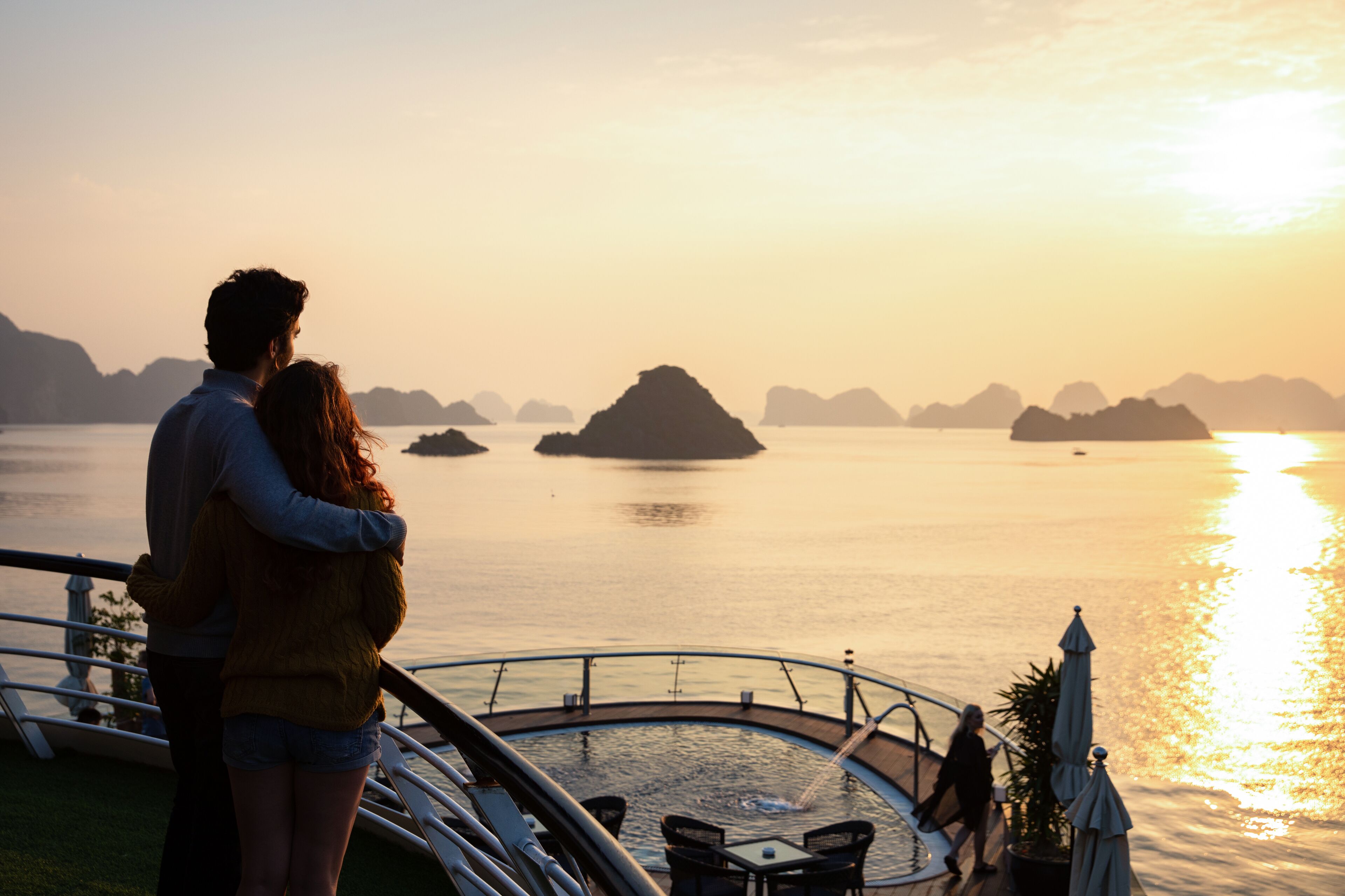 A couple standing on a boat deck overlooking calm waters with distant islands during sunset.