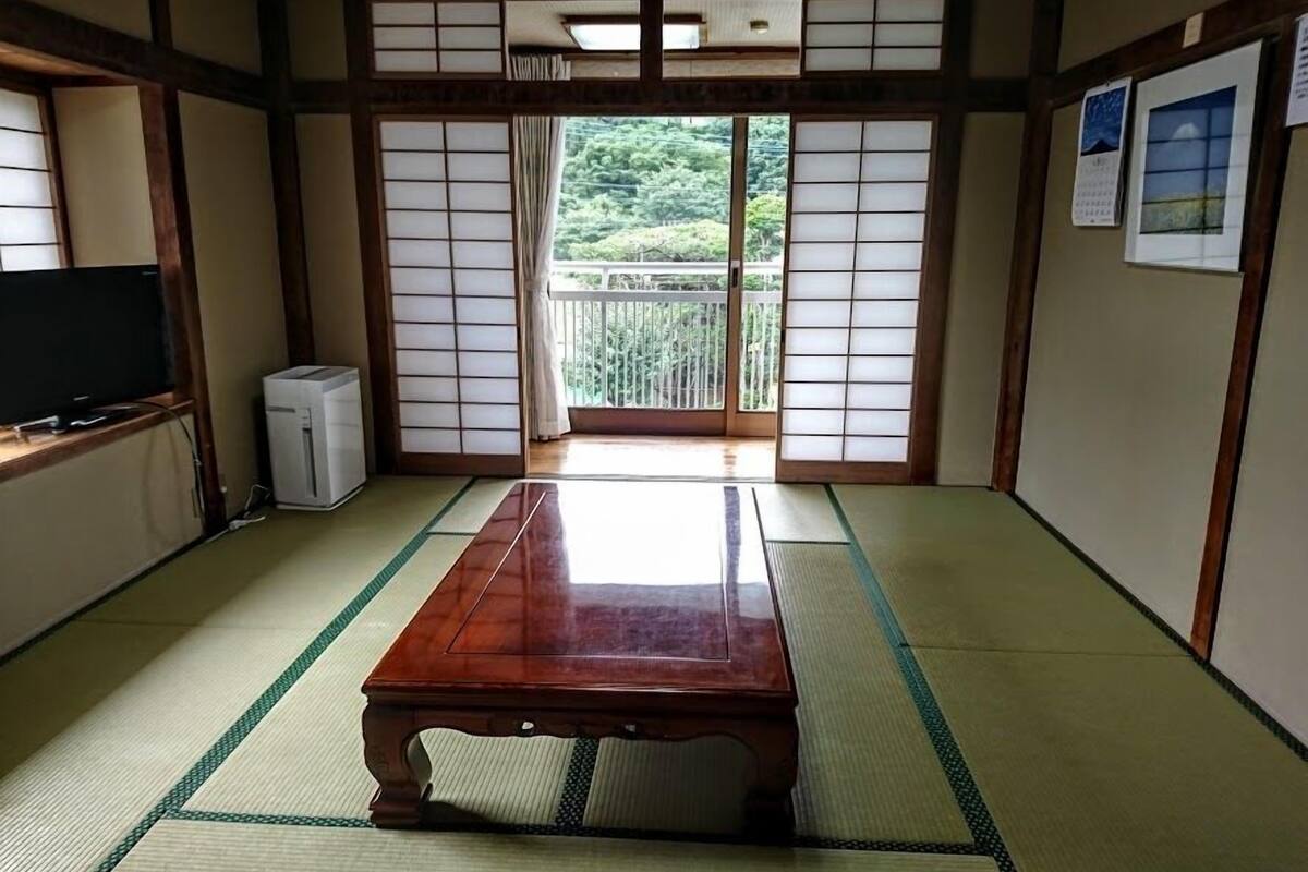 A traditional Japanese room with tatami flooring, a wooden table, a television, and a sliding door leading to a balcony with a view of greenery.
