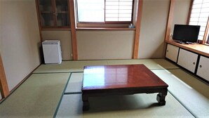Ein traditioneller japanischer Raum mit Tatami-Bodenbelag, einem Holztisch, einem Fernseher auf einem Regal und einem kleinen weißen Gerät.