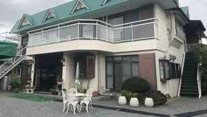 Ein zweistöckiges Gebäude mit einem grünen Dach, einem Balkon mit weißen Geländern und einer überdachten Außenbereich-Sitzgelegenheit mit weißen Stühlen und Tischen.