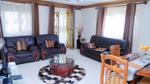 A modern living room with two black leather sofas, a glass coffee table, a wooden dining table with chairs, and a decorative vase with flowers. - Floben Apartments (Kampala)