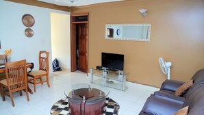 A living room with a round coffee table, a sofa, a TV, and a dining area with chairs and a table. - Floben Apartments (Kampala)